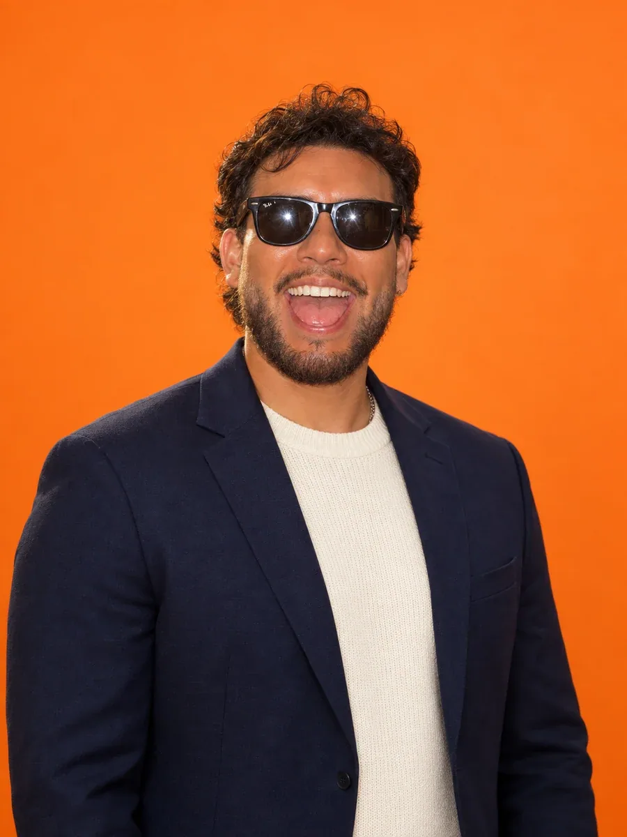 Zivan Zuniga, designer at Zavila Studio, wearing sunglasses and a midnight-blue blazer against an orange backdrop
